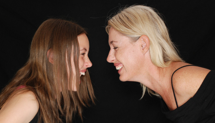 brunette teenager smiling and facing blond woman smiling against black background