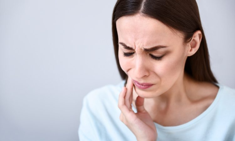 A brunette woman wearing a blue shirt touches her cheek due to a toothache and cringes in pain